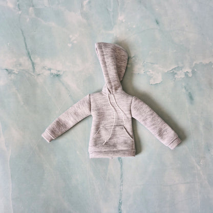 Flat lay of a grey miniature doll-sized hoodie featuring a front kangaroo pocket and white hood drawstrings.