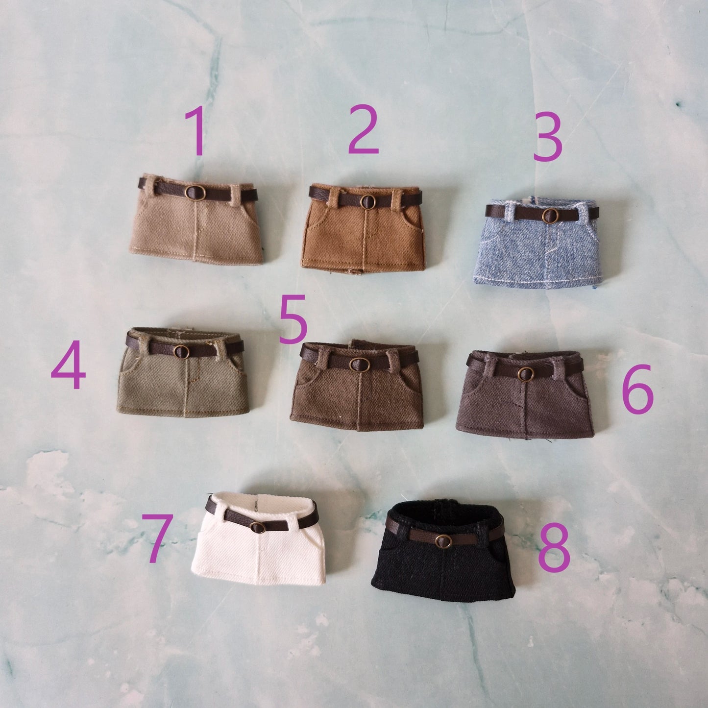 Collection of 8 miniature doll mini skirts with belts, available in beige, camel, blue denim, olive green, dark brown, grey, white, and black.