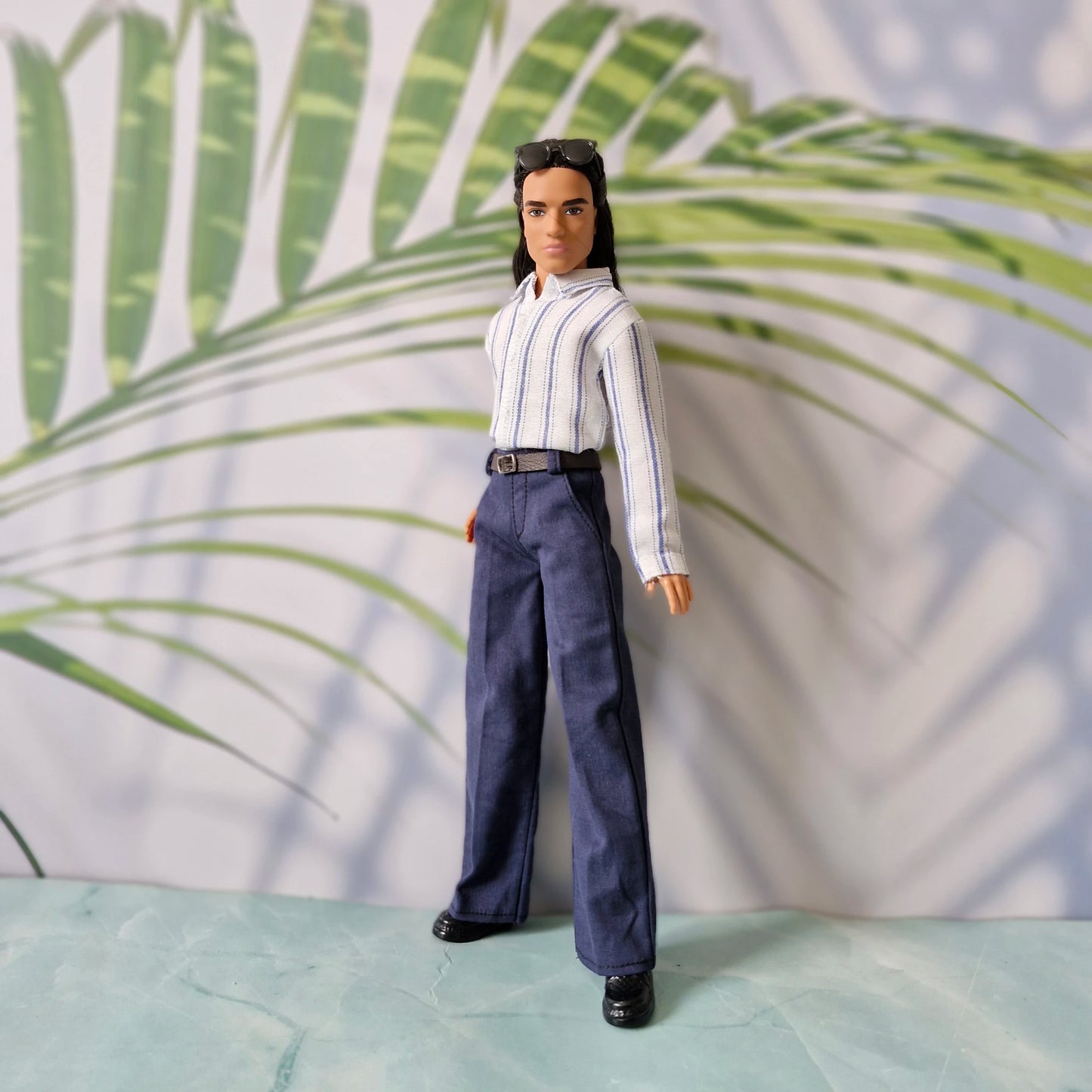 Full body view of a Ken doll in a stylish striped shirt and dark blue slacks, posing against a tropical palm leaf background.