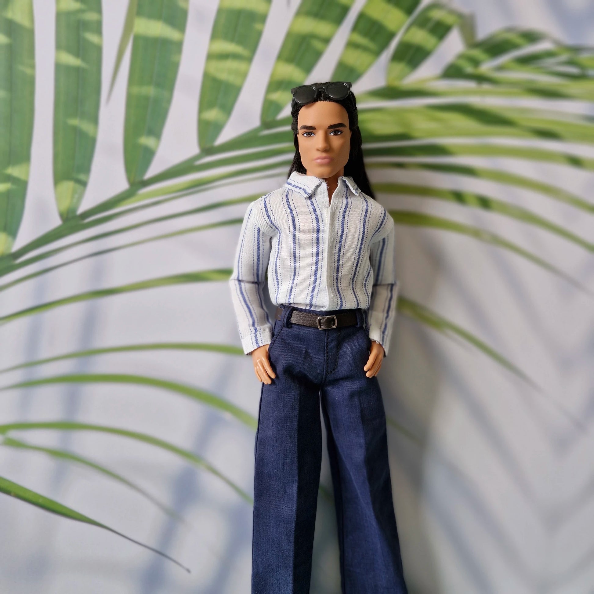 Ken doll dressed in a smart striped collared shirt and blue tailored pants, wearing sunglasses on his head.