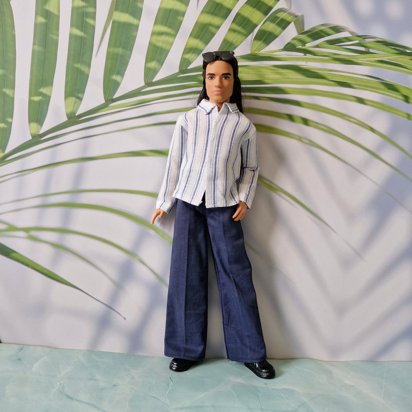 Ken doll modeling a casual untucked blue and white striped shirt with dark navy pants and black shoes.