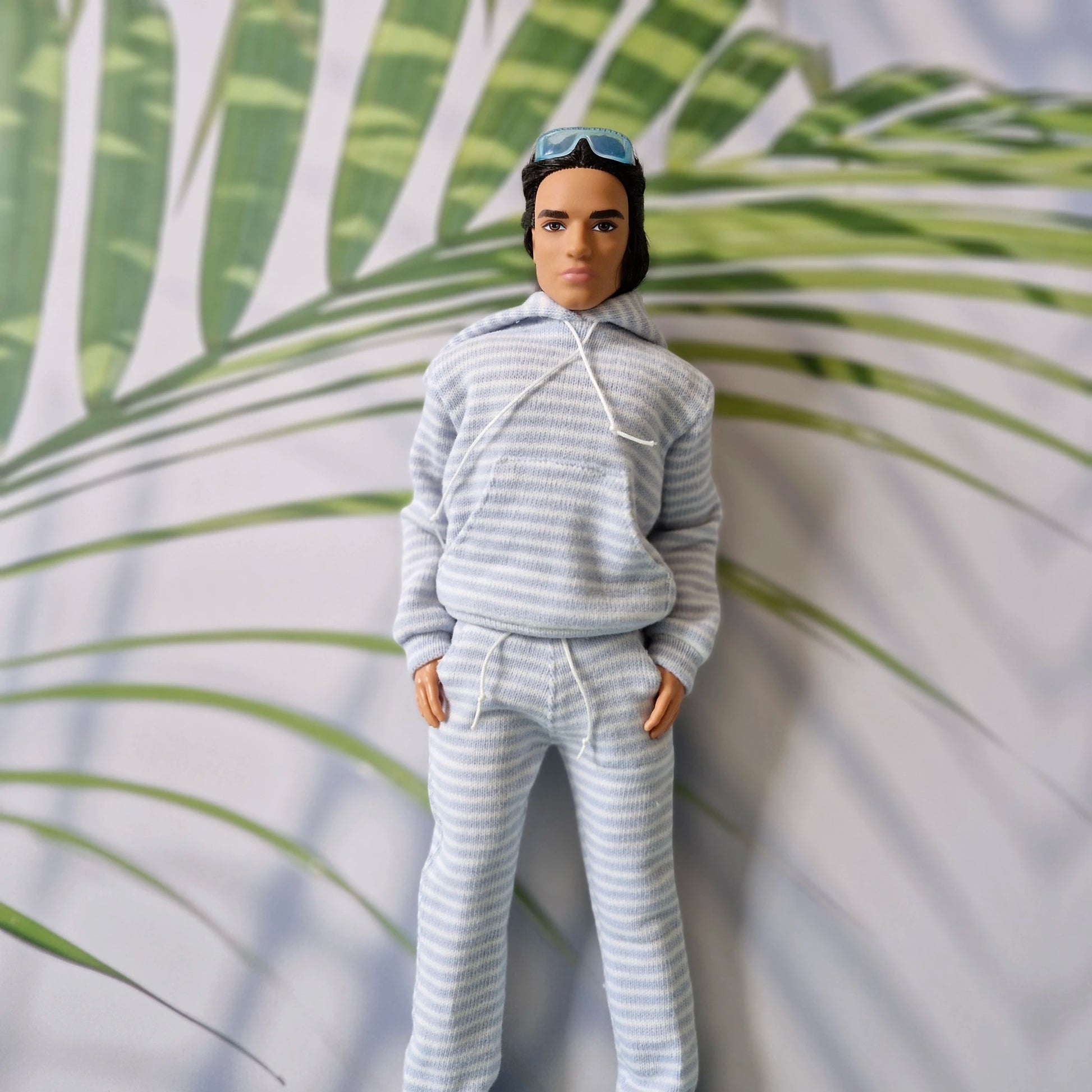 Ken doll wearing a light blue and white striped hoodie with matching tapered sweatpants, standing against a palm leaf background.