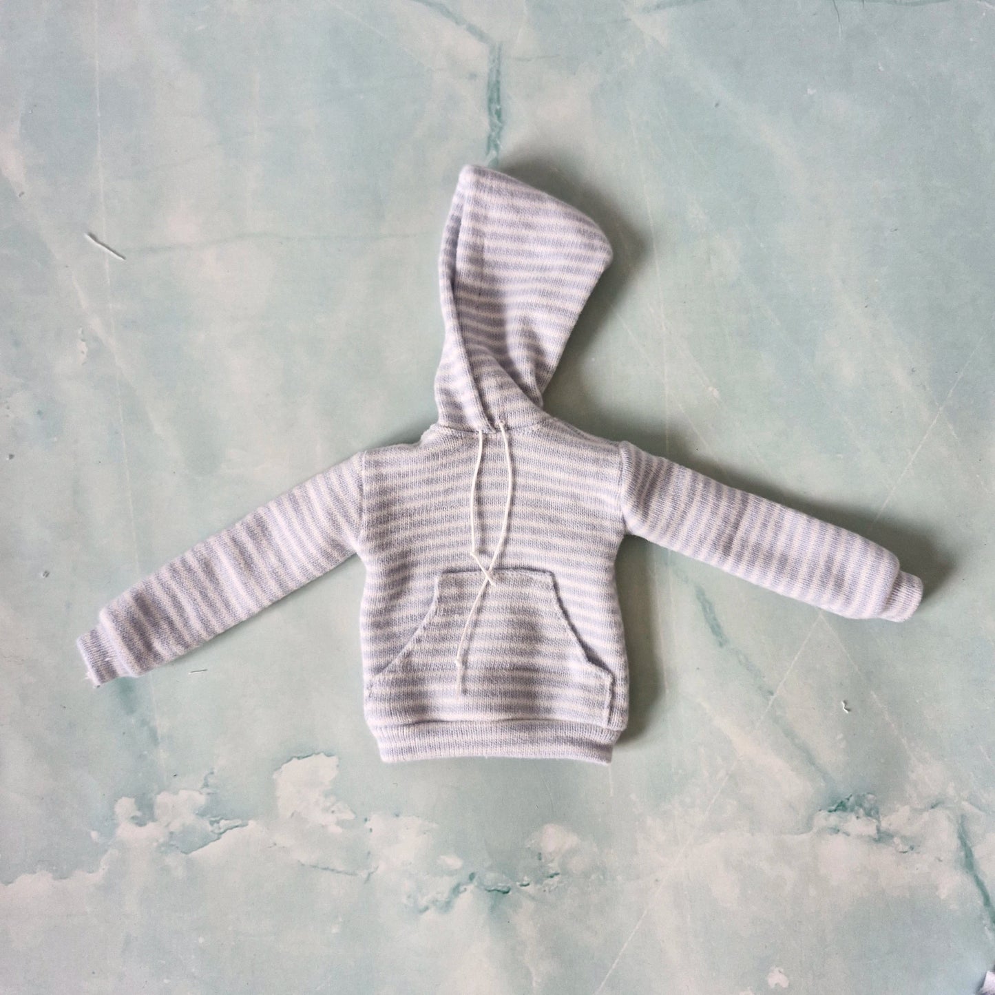 Flat lay of a miniature doll-sized light blue striped hoodie with a front pocket and white drawstrings.