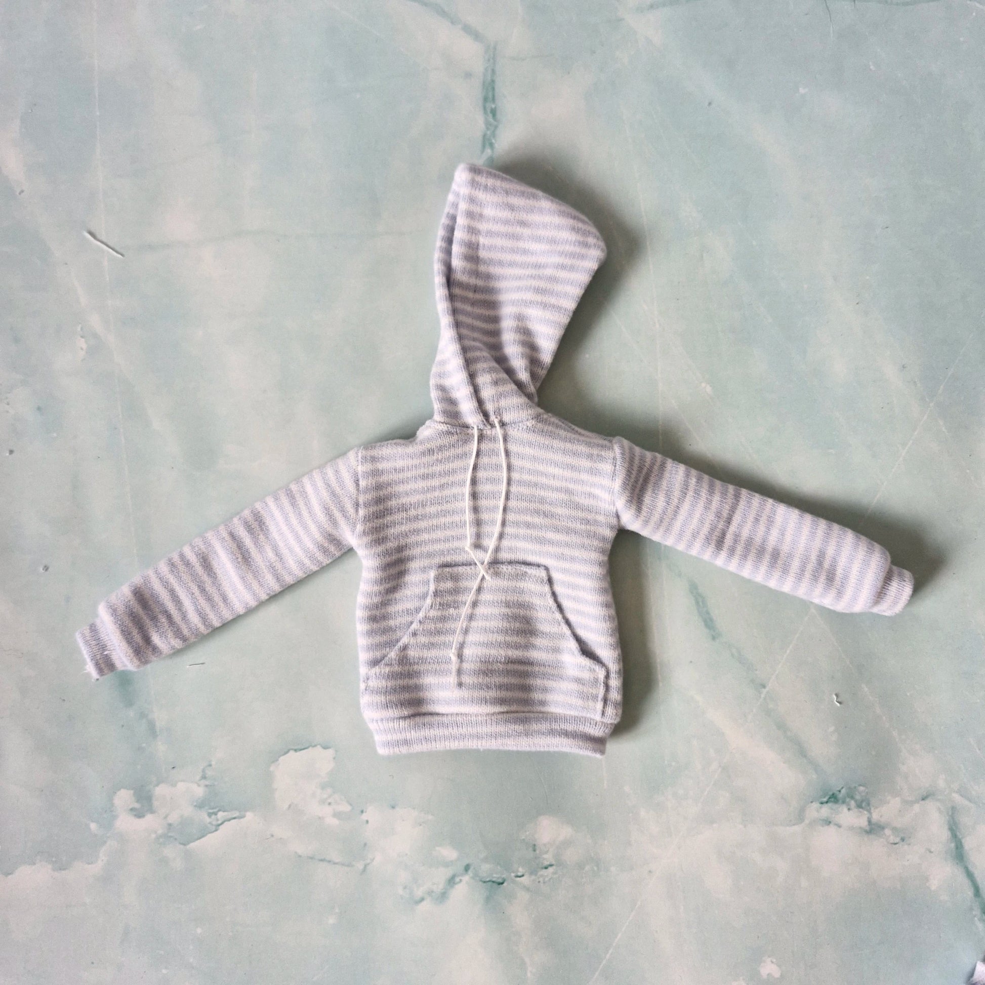 Flat lay of a miniature doll-sized light blue striped hoodie with a front pocket and white drawstrings.