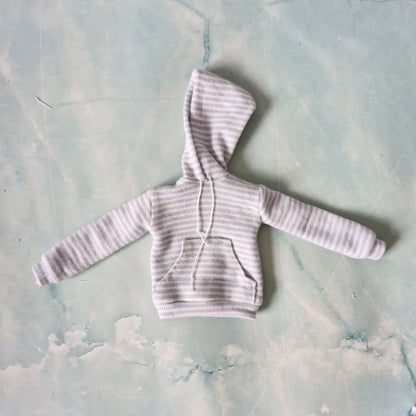 Flat lay of a miniature doll-sized light blue striped hoodie with a front pocket and white drawstrings.