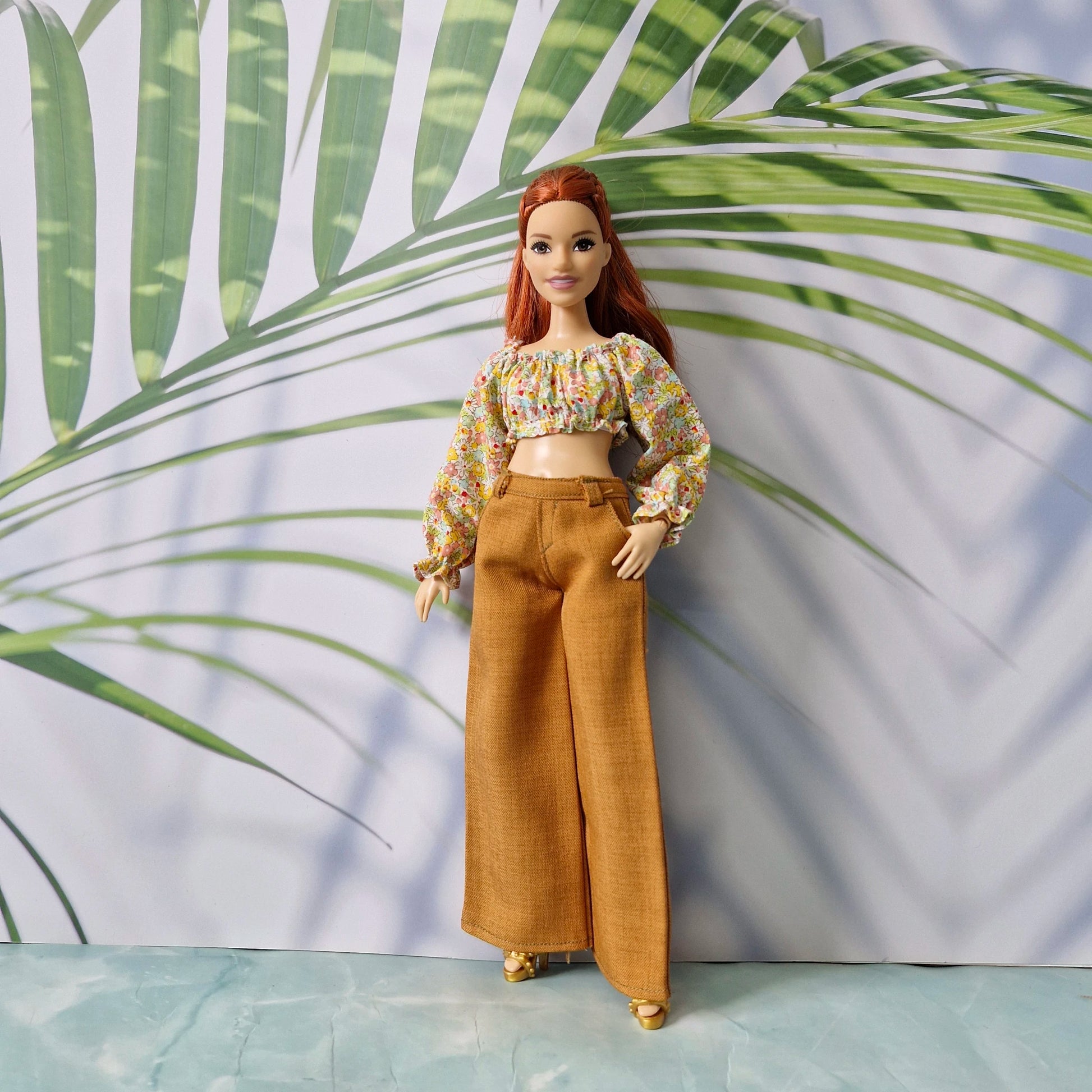 Redhead Barbie doll wearing a floral off-shoulder long-sleeve crop top and brown wide-leg pants against a tropical leaf background.