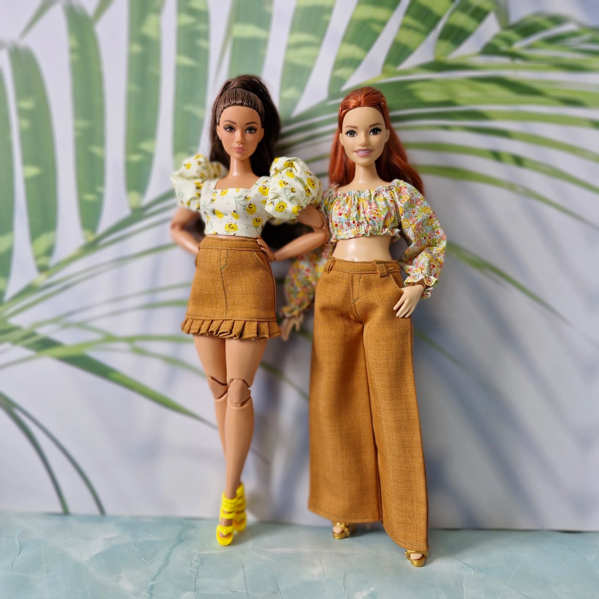 Two Barbie dolls showcasing summer fashion: one in a floral puff-sleeve top and brown ruffled skirt, the other in an off-shoulder top and wide-leg pants.