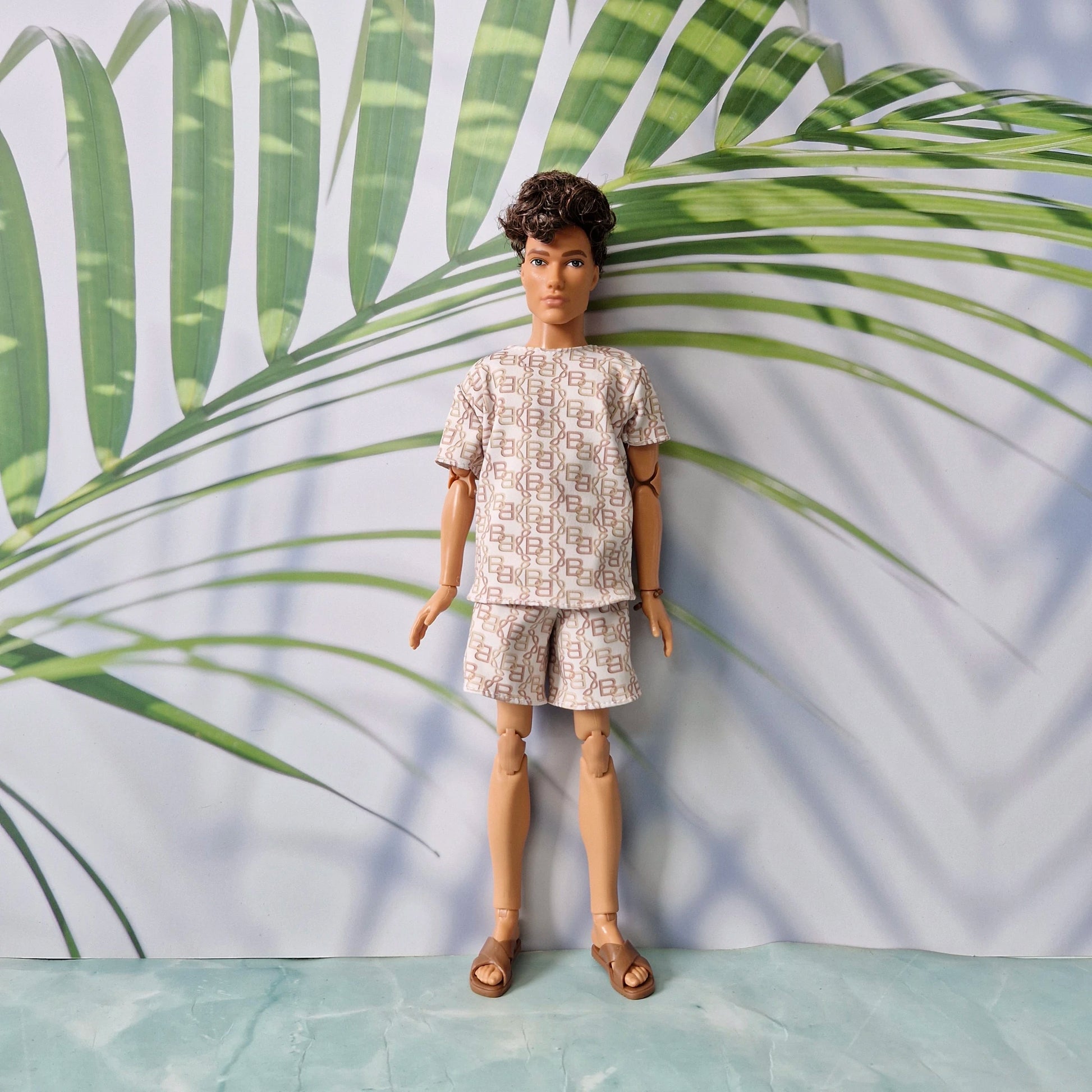 A fashion doll with short dark hair stands wearing a matching short-sleeve top and shorts set in a beige and white geometric pattern, paired with brown sandals, against a backdrop with a palm leaf print.