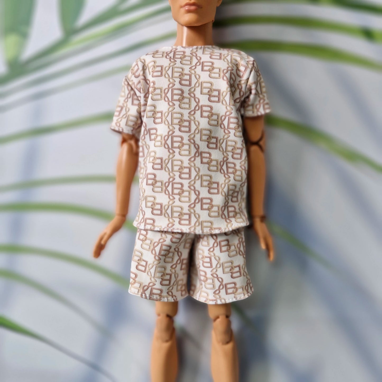 A doll or mannequin wearing a short-sleeve matching set with an allover geometric monogram-style print in brown and beige on a light background, posed against a softly blurred leafy backdrop.