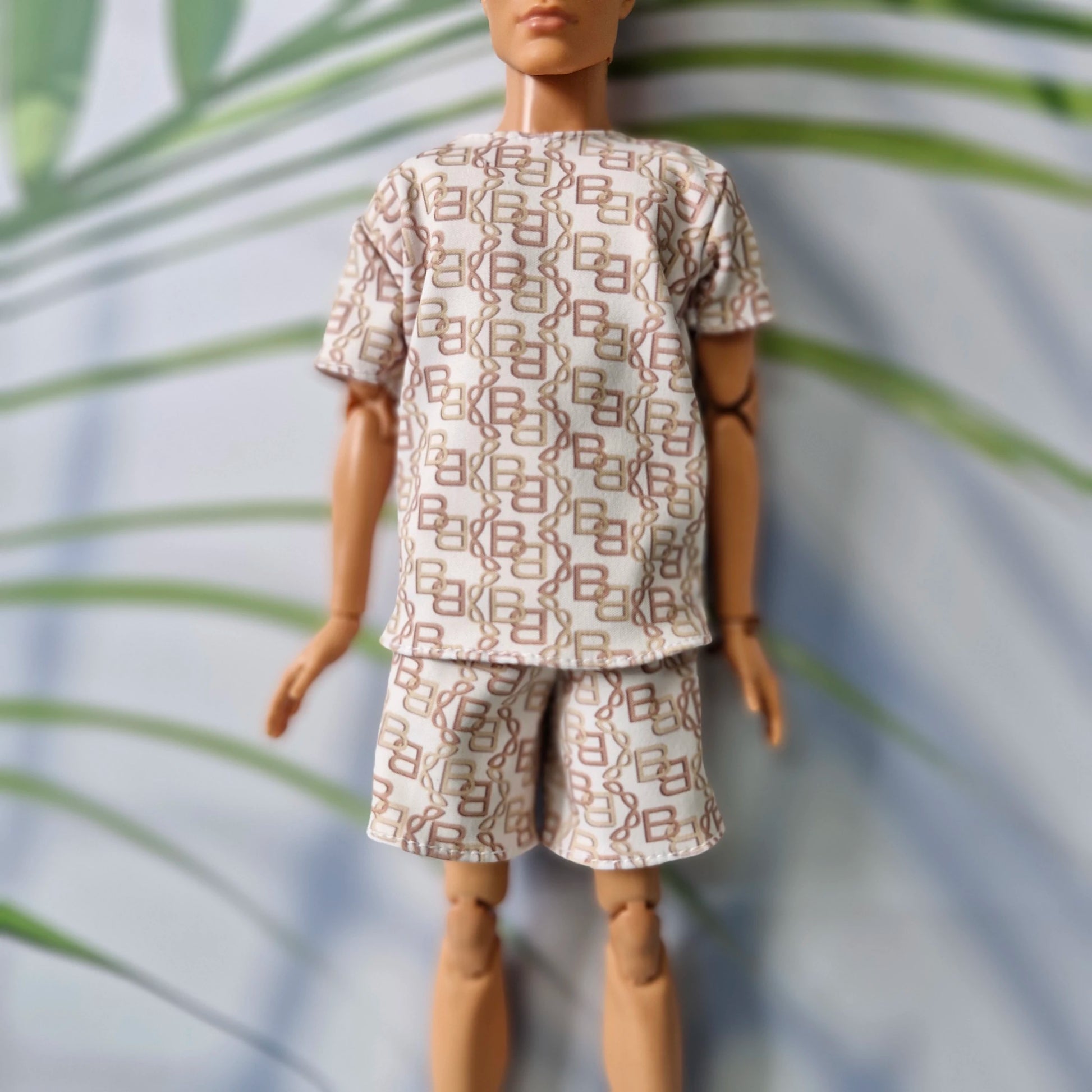 A doll or mannequin wearing a short-sleeve matching set with an allover geometric monogram-style print in brown and beige on a light background, posed against a softly blurred leafy backdrop.