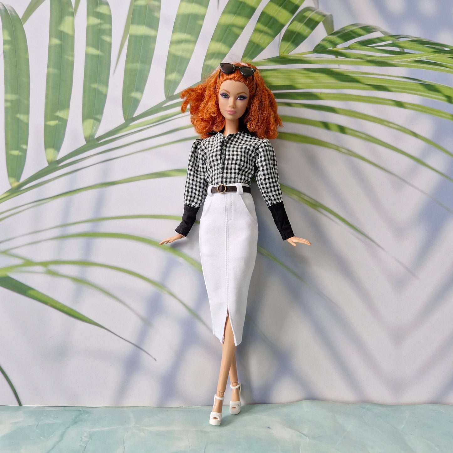 Full body shot of a doll modeling trendy clothes for Barbie: a gingham blouse and a tailored white skirt with a slit.