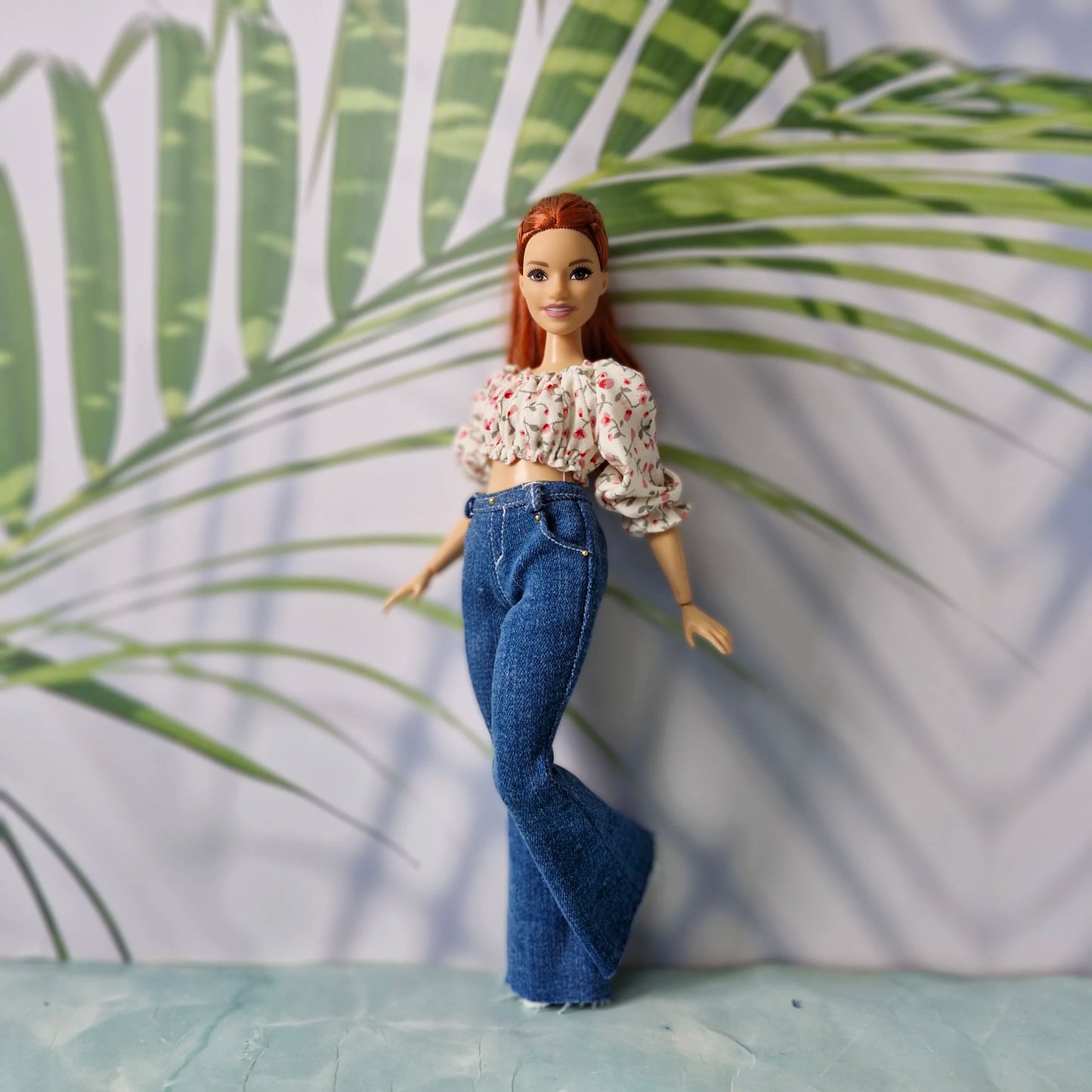 Redhead curvy doll wearing a pink floral crop top and dark blue flared jeans for Curvy Barbie, posing with one leg up.