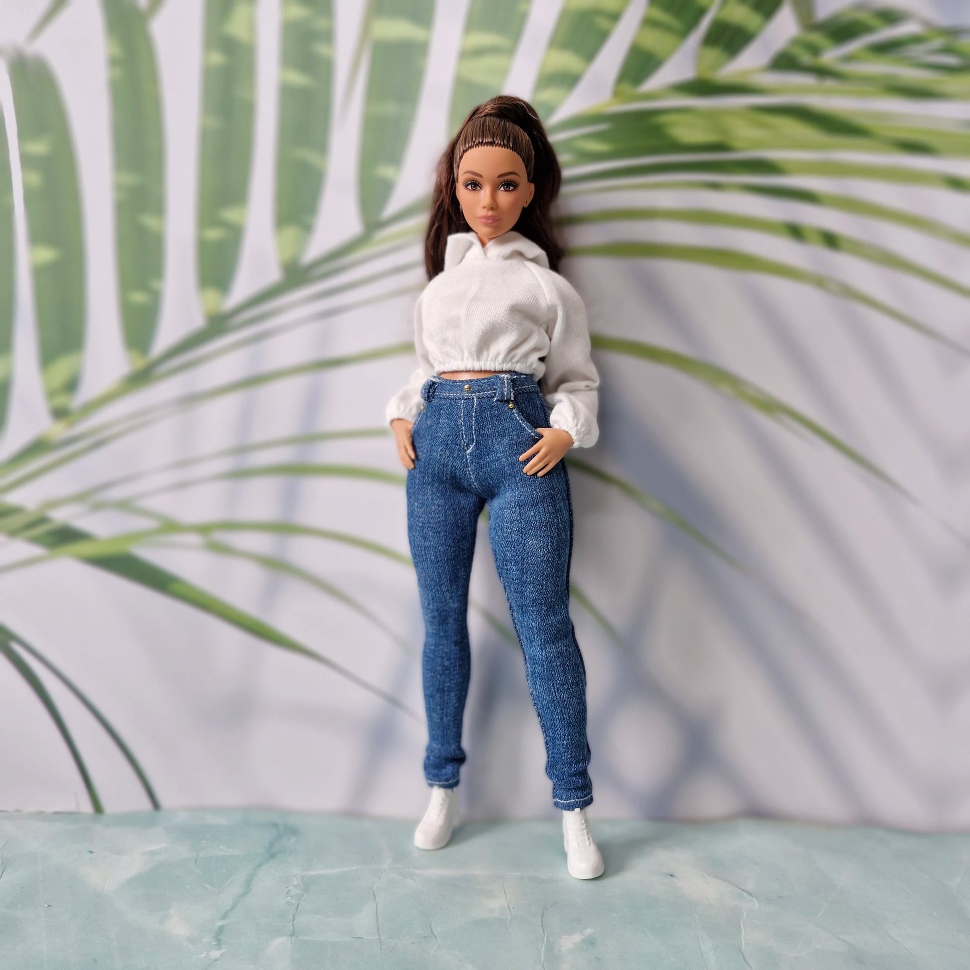 Brunette fashion doll with hands in pockets wearing a white hoodie and dark wash skinny jeans for Curvy Barbie.