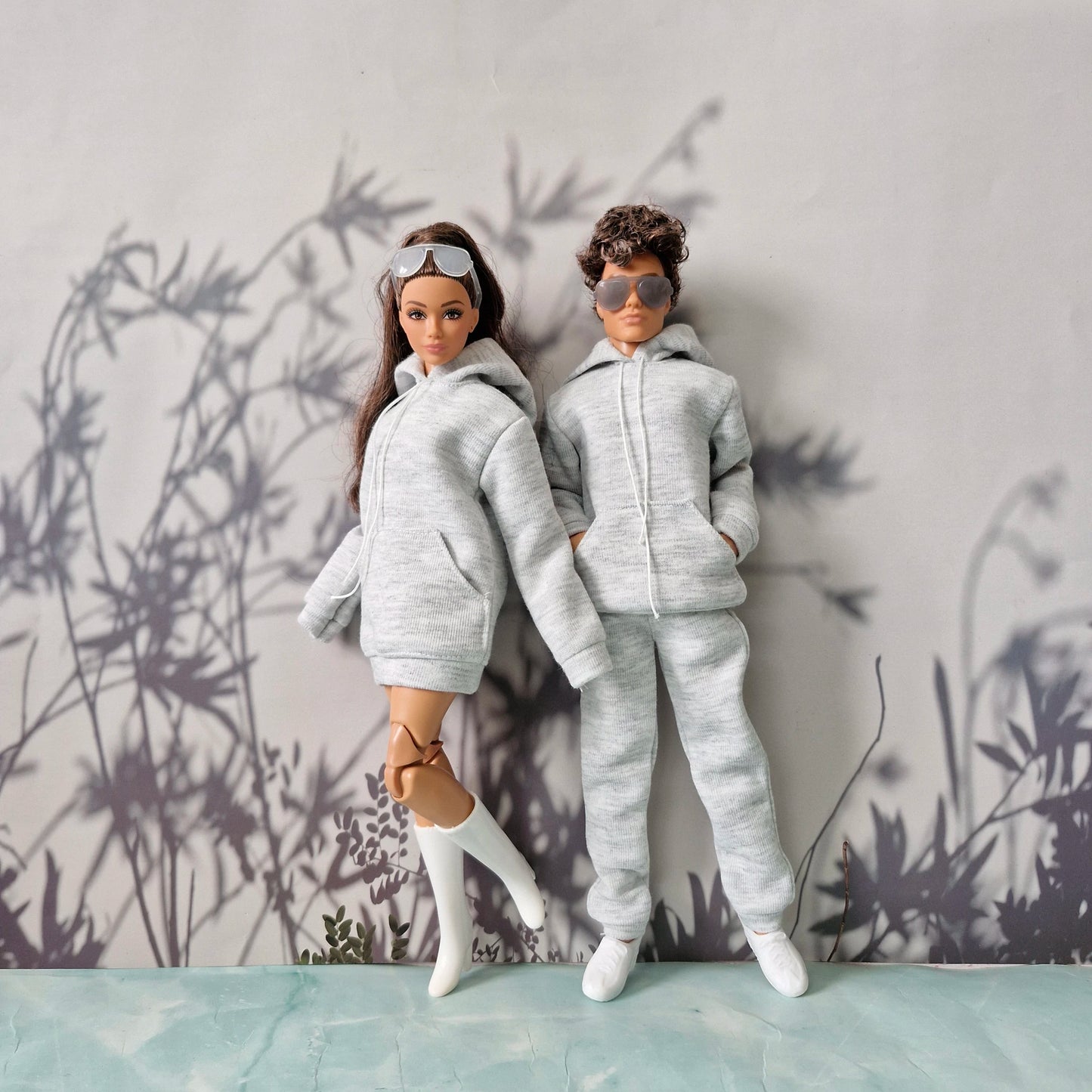 Two fashion dolls dressed in matching light gray hoodies and sweatpants sets, standing against a gray wall with a leafy shadow pattern and a pale blue surface below.