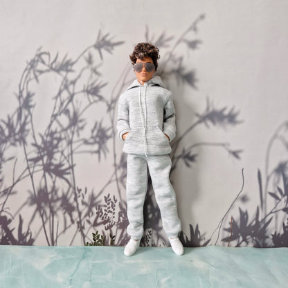A doll or miniature figure wearing a light gray heather zip-up hoodie and matching sweatpants, with white sneakers and dark sunglasses, standing against a gray wall with a bamboo leaf shadow pattern.