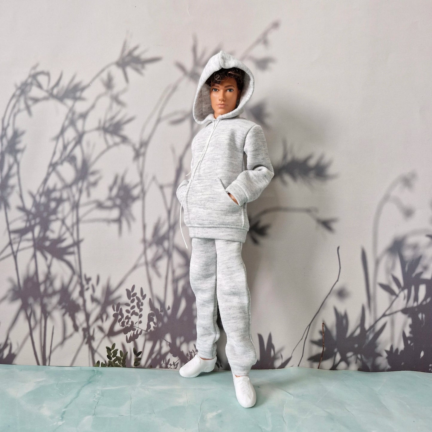A doll or mannequin wearing a light gray heathered hooded sweatshirt and matching jogger pants, standing against a pale wall with dark botanical shadows and a mint-green surface below.