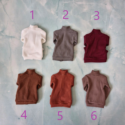 Six miniature ribbed knit turtleneck sweaters for dolls, numbered 1 to 6 in purple text, arranged on a light blue marbled background.