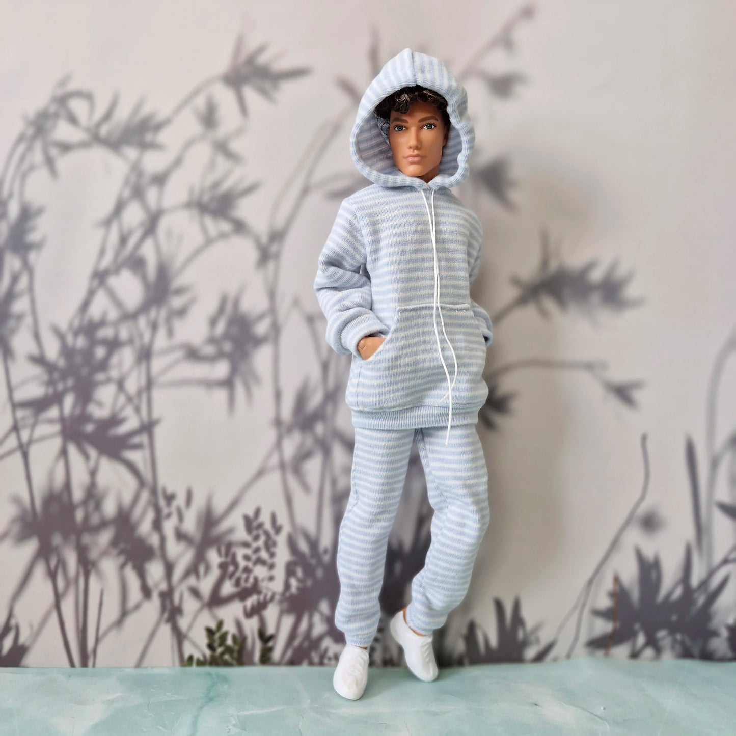 A doll or mannequin wearing a light blue and white striped hooded sweatshirt and matching pants, standing against a gray wall with bamboo silhouettes.