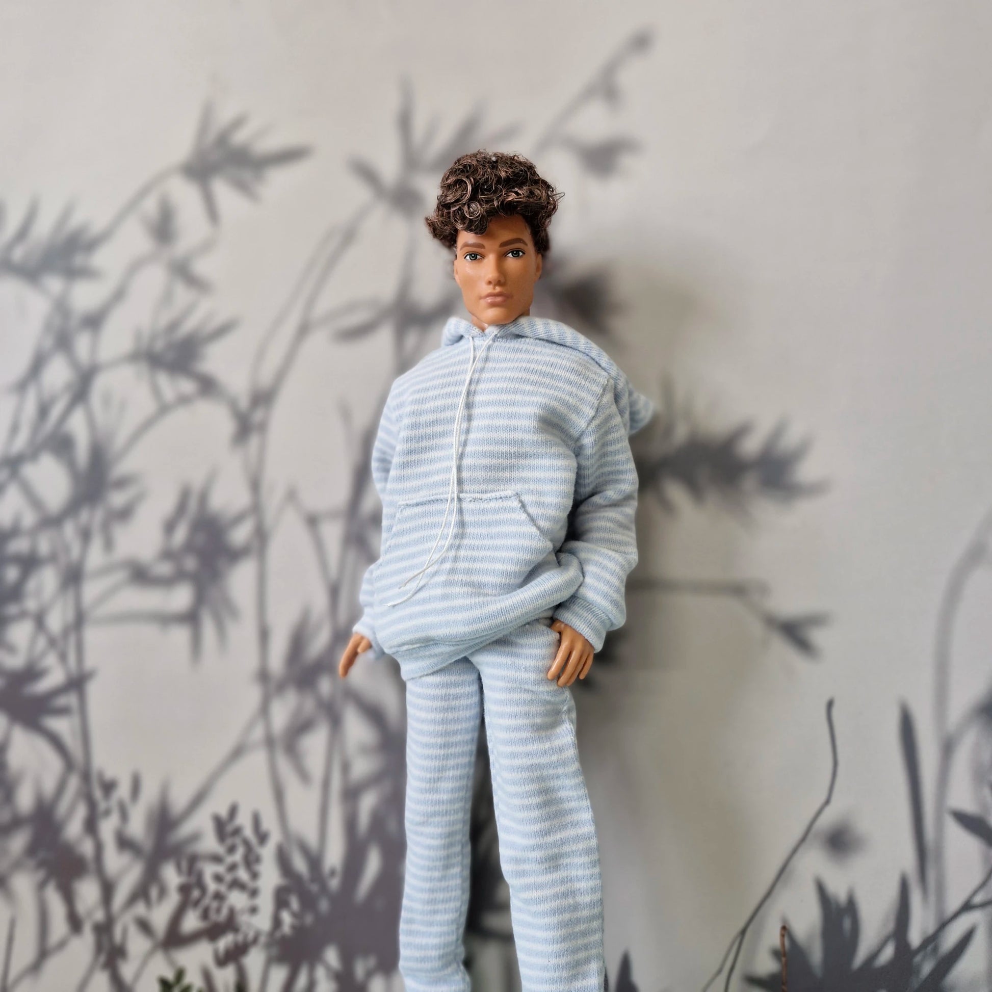 A doll with curly brown hair wearing a light blue and white striped hooded sweatshirt and matching pants, posed against a gray wall with black leaf shadows.