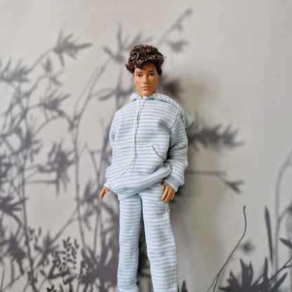 A doll with curly brown hair wearing a light blue and white striped hooded sweatshirt and matching pants, posed against a gray wall with black leaf shadows.