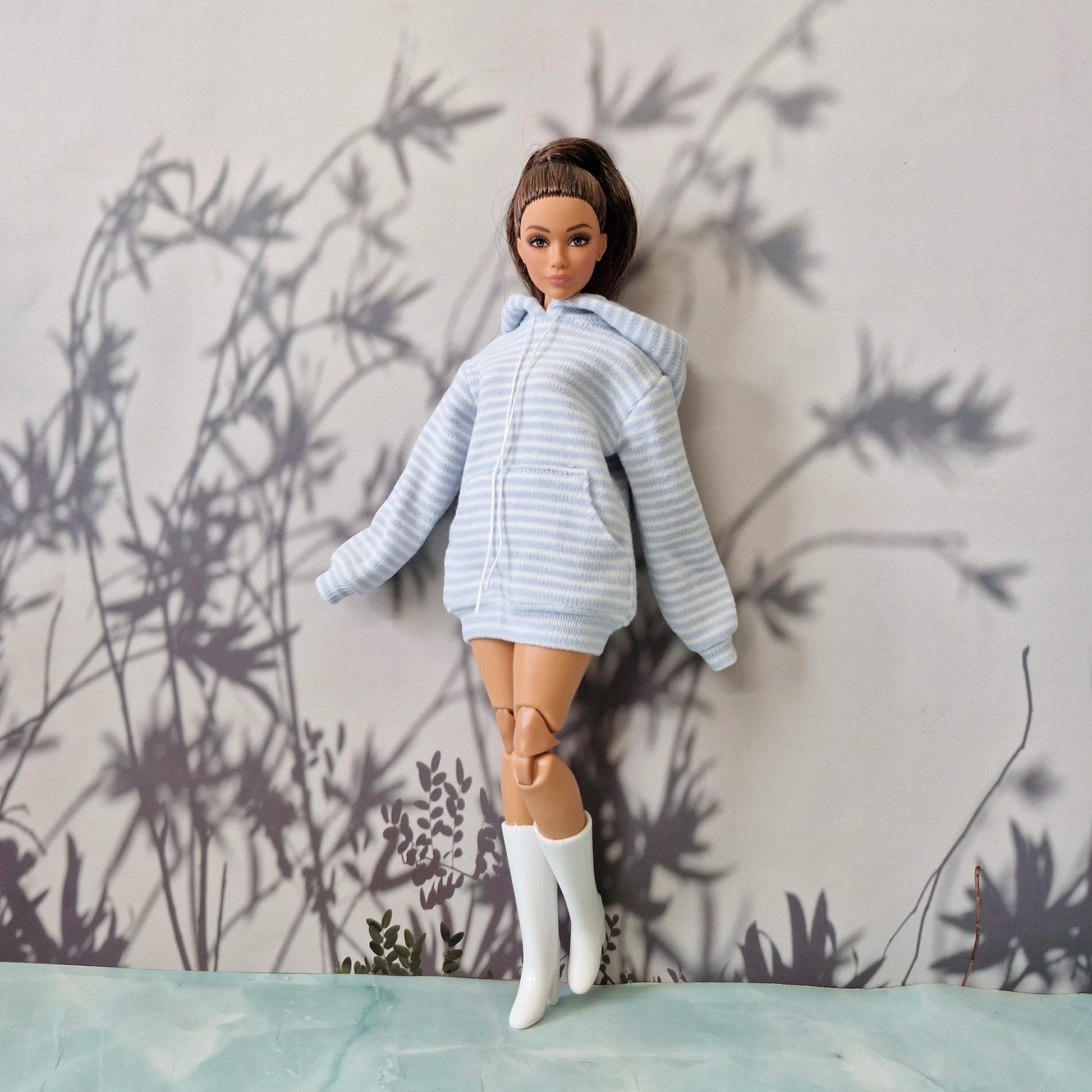 A doll with brown hair in a ponytail wearing a light blue and white striped zip-up hoodie, a short skirt, and white boots, posed against a gray wall with leaf shadows.