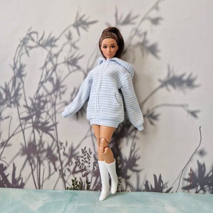 A doll with brown hair in a ponytail wearing a light blue and white striped zip-up hoodie, a short skirt, and white boots, posed against a gray wall with leaf shadows.