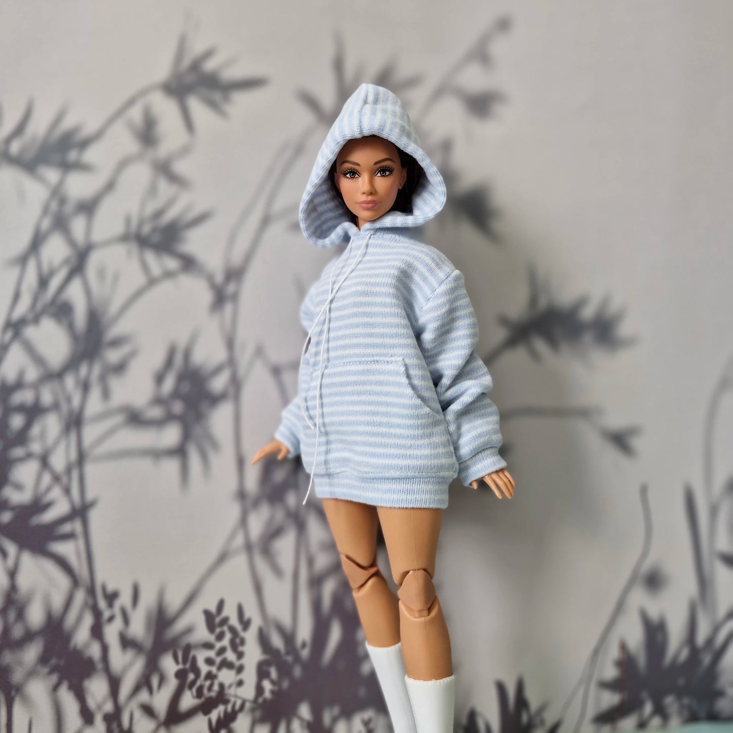 A doll with brown articulated legs and a striped light blue and white hooded sweatshirt dress, standing against a gray wall with black leaf shadows.
