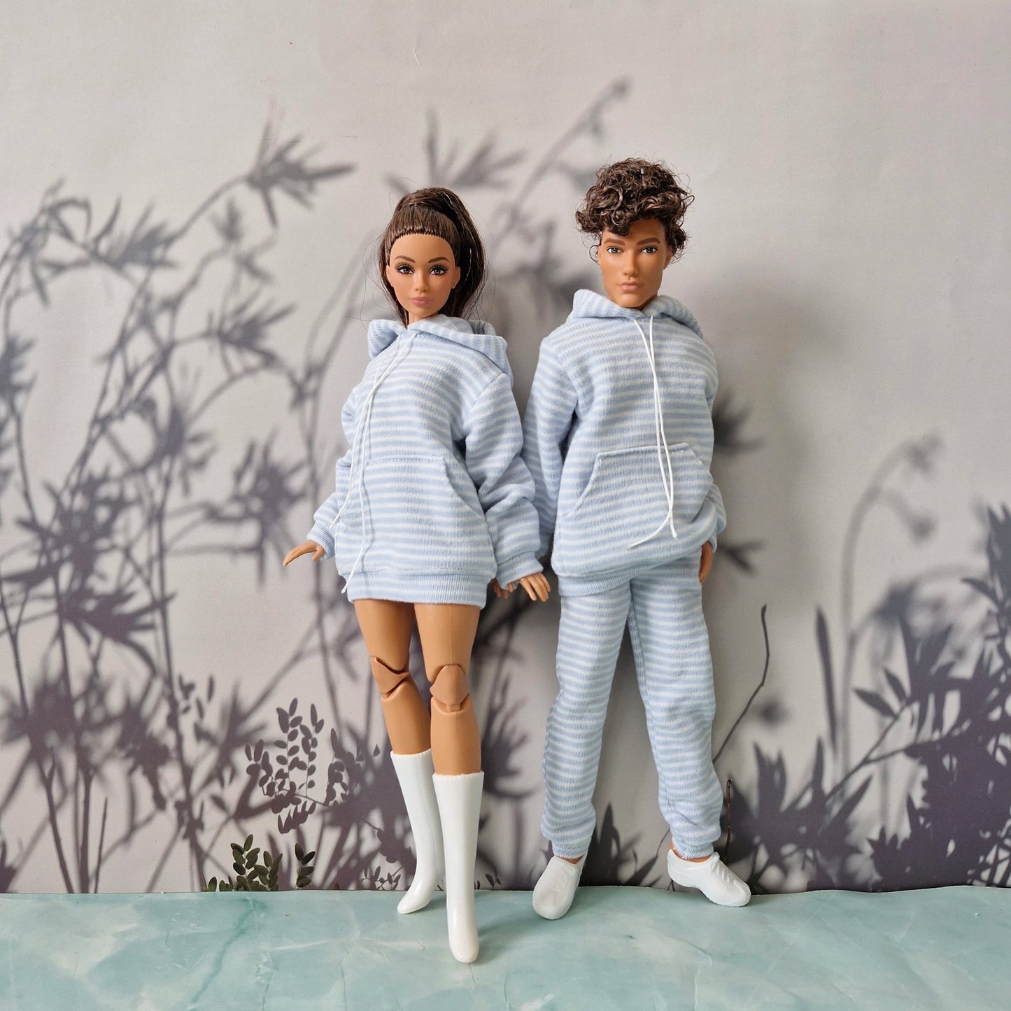 Two fashion dolls wearing matching light blue and white striped hoodies and joggers, standing against a gray wall with a bamboo shadow pattern.