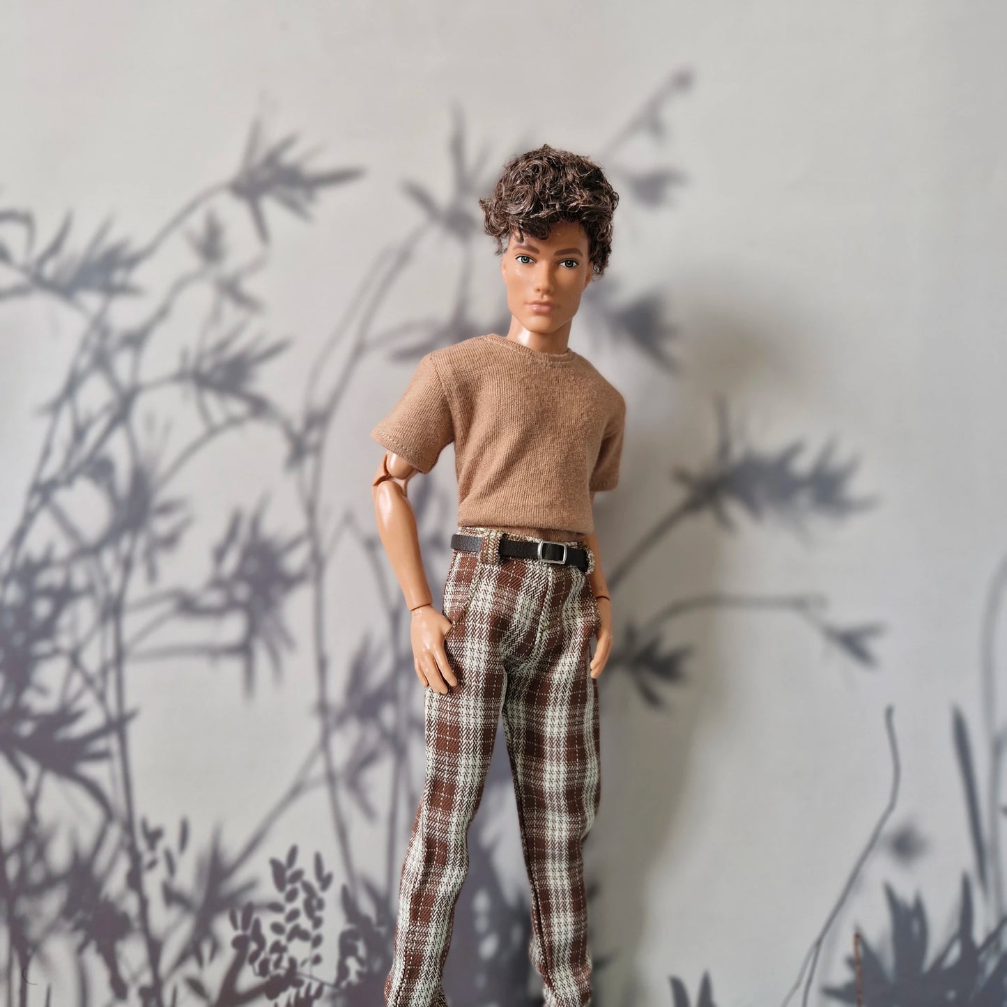 A doll with short curly brown hair stands against a light gray wall with dark leaf shadows. The doll is wearing a tan short-sleeve knit top and brown-and-white plaid pants with a black belt.