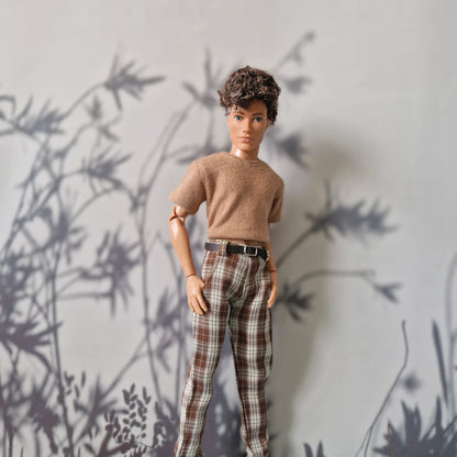 A doll with short curly brown hair stands against a light gray wall with dark leaf shadows. The doll is wearing a tan short-sleeve knit top and brown-and-white plaid pants with a black belt.