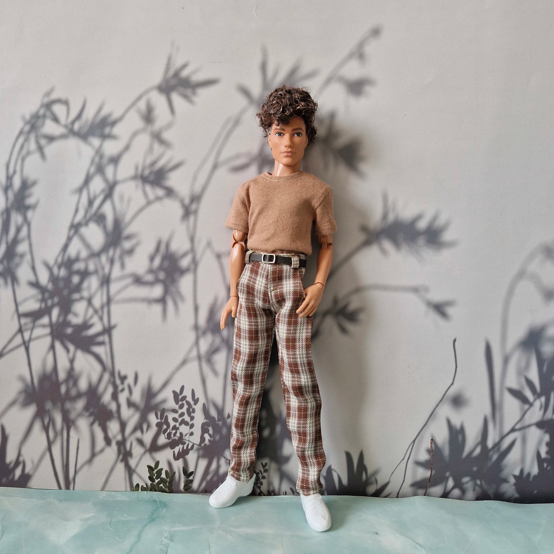 A doll or fashion mannequin with short curly brown hair stands wearing a beige short-sleeve top, brown and beige plaid pants with a black belt, and white shoes, posed against a gray wall with black leafy shadow graphics and a pale blue surface below.