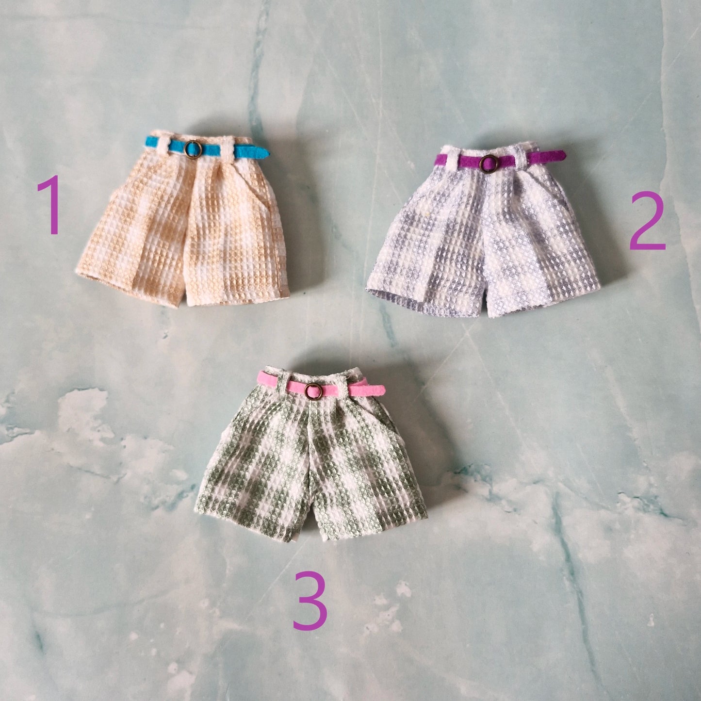 Flat lay of three 1/6 scale doll plaid shorts in beige, blue, and green sold separately for Barbie.