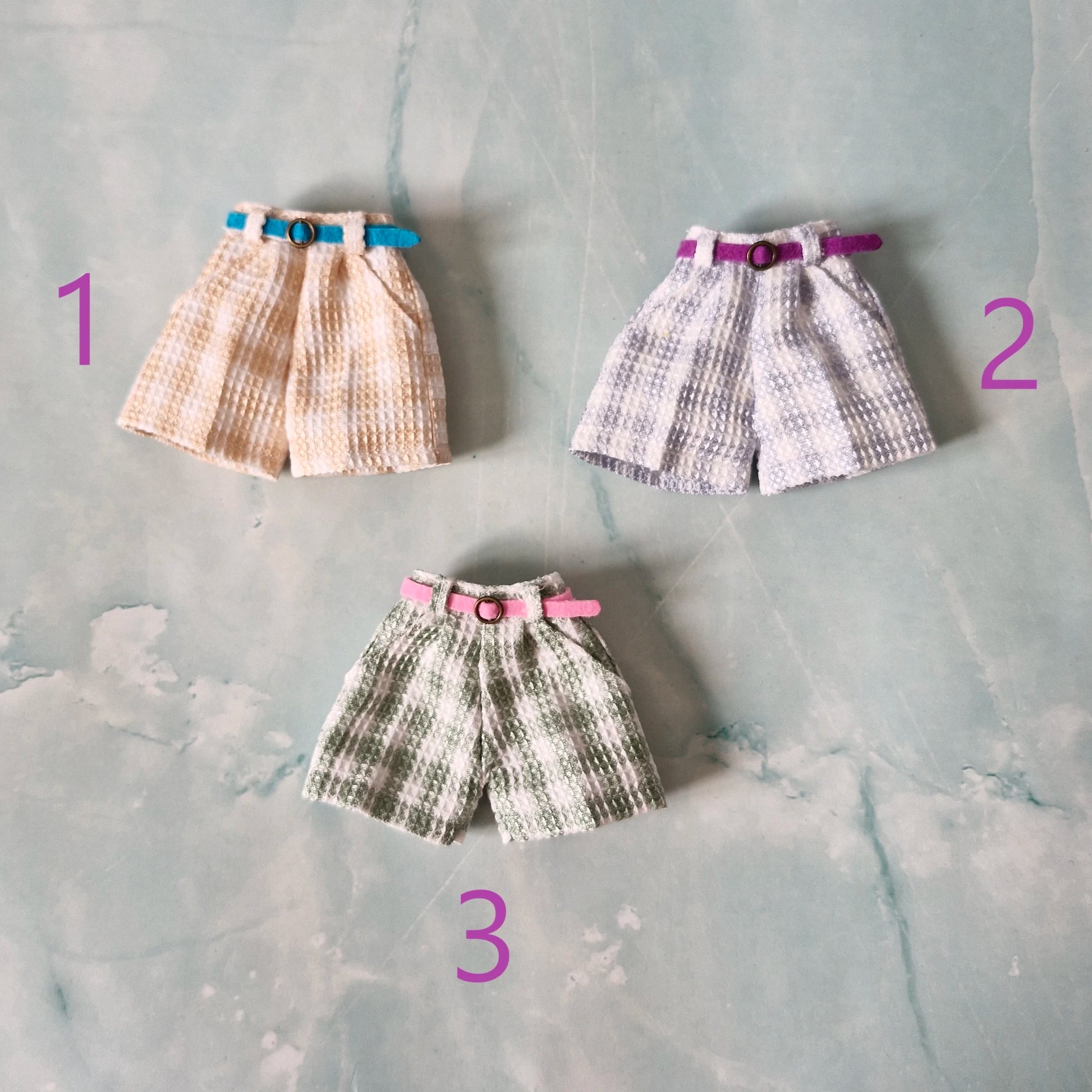 Flat lay of three 1/6 scale doll plaid shorts in beige, blue, and green sold separately for Barbie.