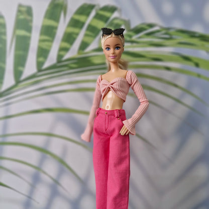 A fashion doll with blonde hair and blue eyes, wearing a pink ribbed knit crop top with long sleeves and matching pink high-waisted wide-leg jeans, standing against a background with palm leaf shadows.