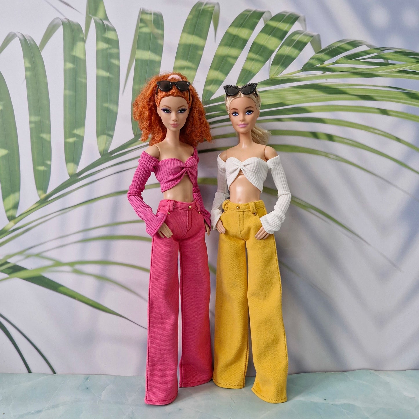Two fashion dolls standing side by side in front of a tropical leaf backdrop. The doll on the left has curly orange hair and wears a bright pink off-the-shoulder crop top with matching wide-leg pants.