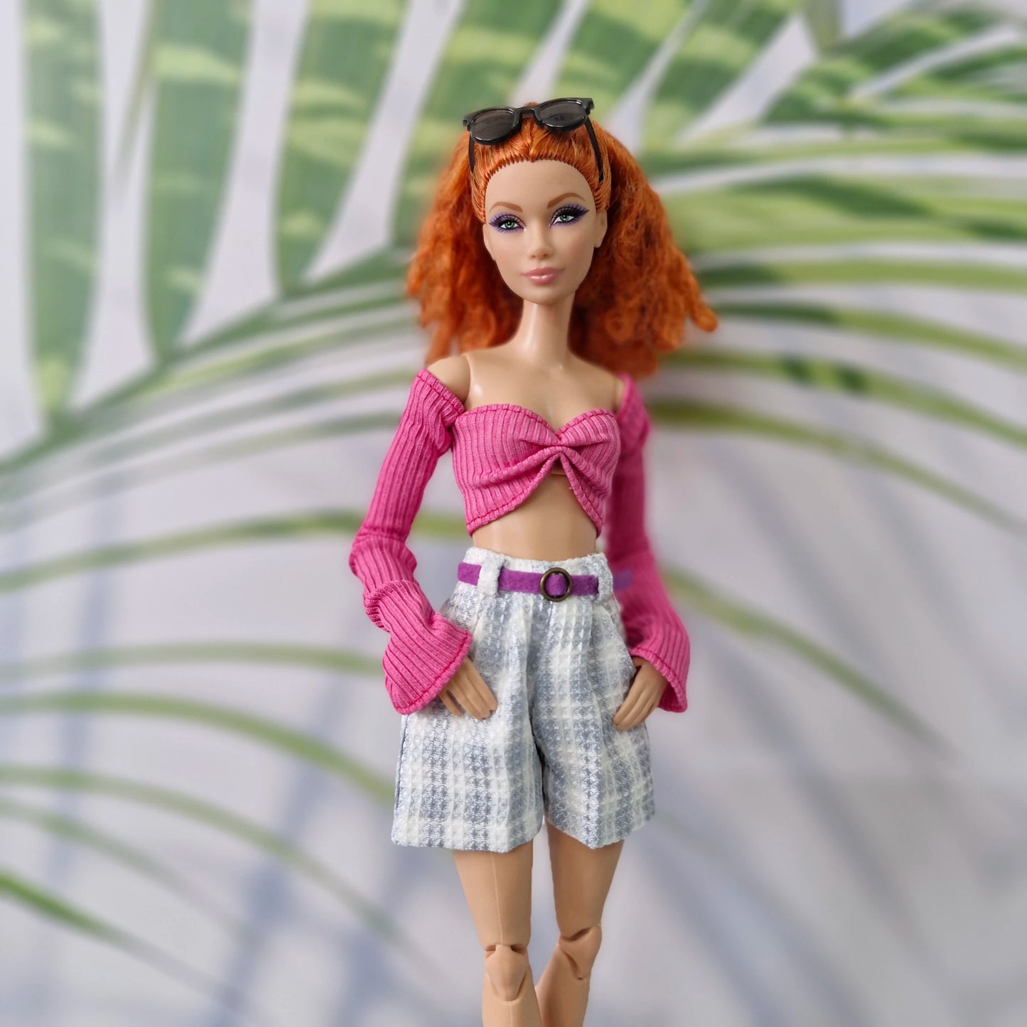 A fashion doll with red hair and sunglasses on her head, wearing a pink knit long-sleeve crop top with a large bow, white and gray patterned shorts, and a purple belt, posed against a background of green palm leaves.
