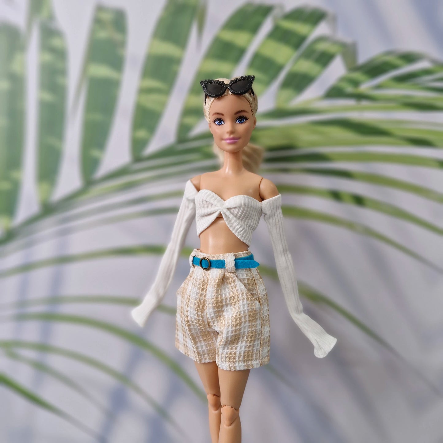 A fashion doll with blonde hair and blue eyes wearing a white ribbed off-the-shoulder crop top with long sleeves, beige-and-white patterned shorts, a bright blue belt, and black sunglasses, posed against a blurred tropical leaf background.
