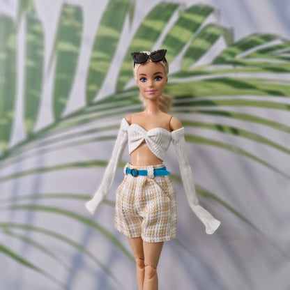 A fashion doll with blonde hair and blue eyes wearing a white ribbed off-the-shoulder crop top with long sleeves, beige-and-white patterned shorts, a bright blue belt, and black sunglasses, posed against a blurred tropical leaf background.