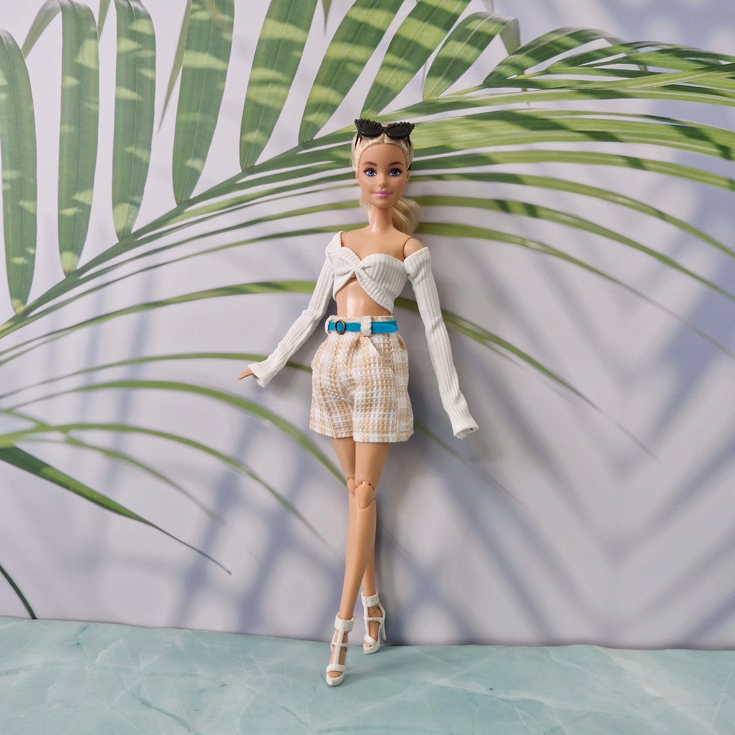 A fashion doll stands against a printed backdrop with large green palm leaves and a pale lavender background. The doll is wearing a white off-the-shoulder long-sleeve crop top, beige-and-white patterned shorts, a bright blue belt, and white high-heeled sandals.