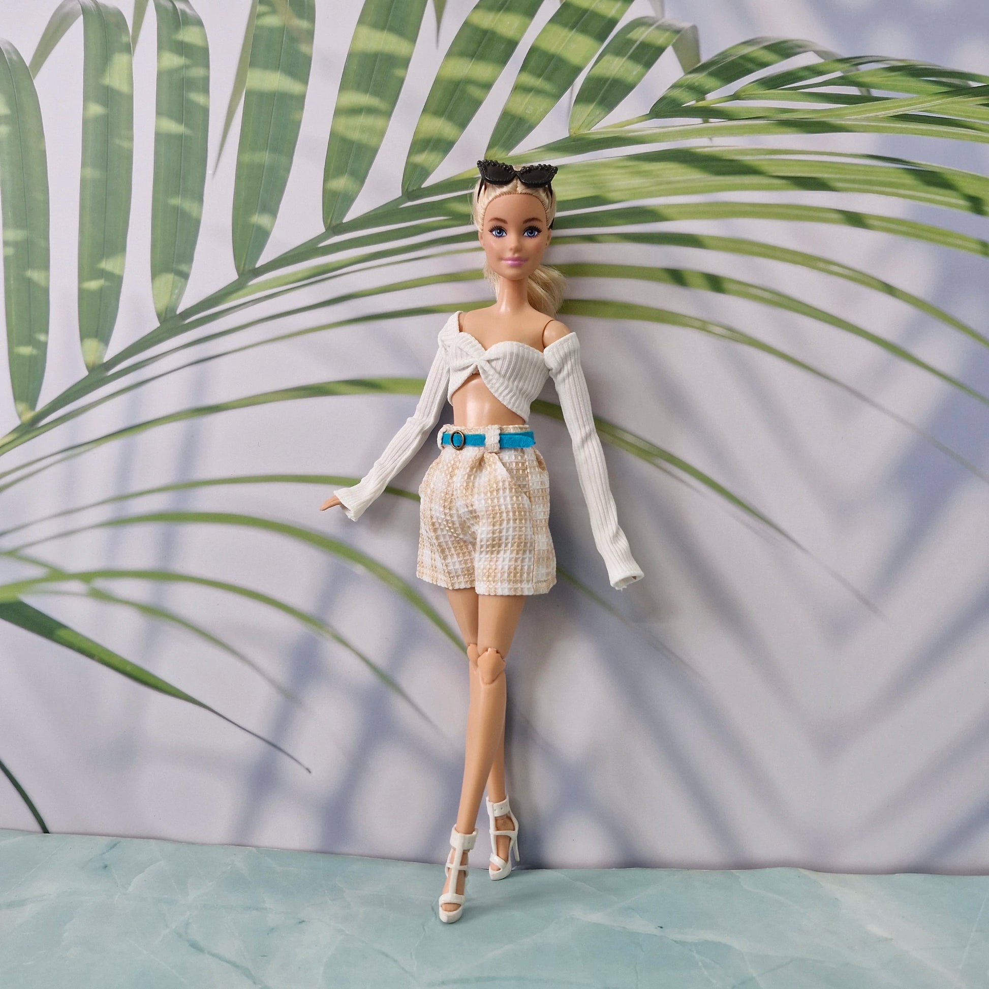 A fashion doll stands against a printed backdrop with large green palm leaves and a pale lavender background. The doll is wearing a white off-the-shoulder long-sleeve crop top, beige-and-white patterned shorts, a bright blue belt, and white high-heeled sandals.