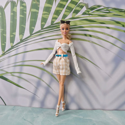 A fashion doll stands against a printed backdrop with large green palm leaves and a pale lavender background. The doll is wearing a white off-the-shoulder long-sleeve crop top, beige-and-white patterned shorts, a bright blue belt, and white high-heeled sandals.