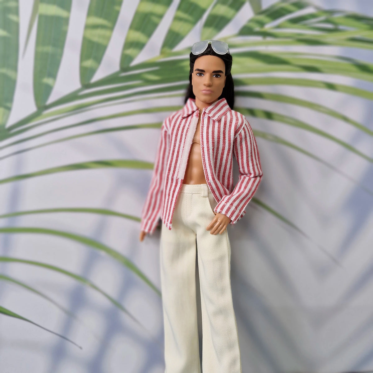 Close-up of a Ken doll in a handmade open red striped collared shirt and white trousers.