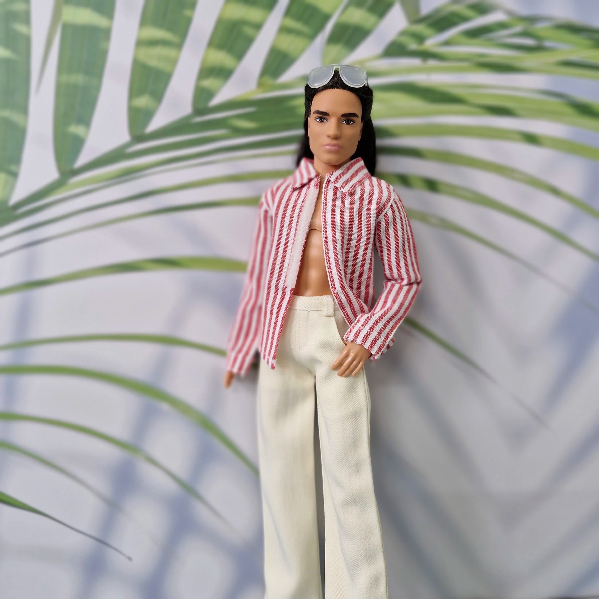 Close-up of a Ken doll in a handmade open red striped collared shirt and white trousers.