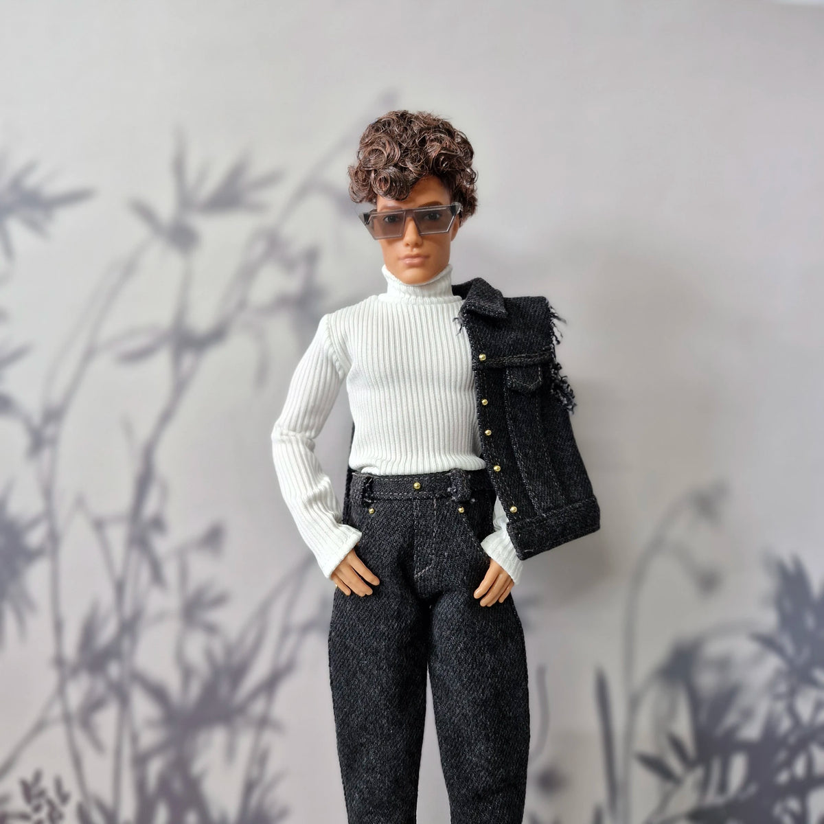 Ken doll modeling a white turtleneck top, dark wash jeans, and a dark denim vest slung over his shoulder.