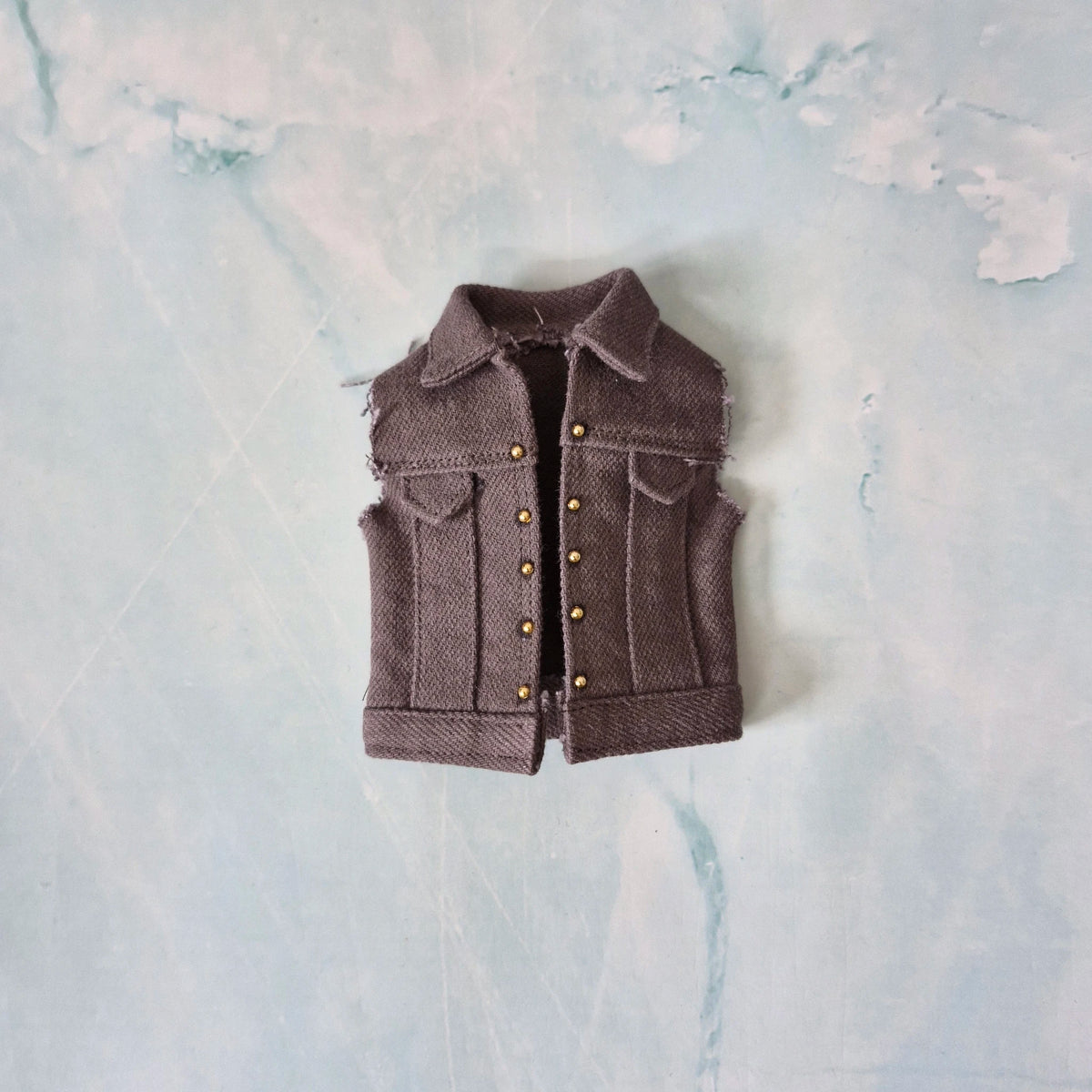 Flat lay of a custom handmade grey denim vest with gold stud details for 1/6 scale male dolls.