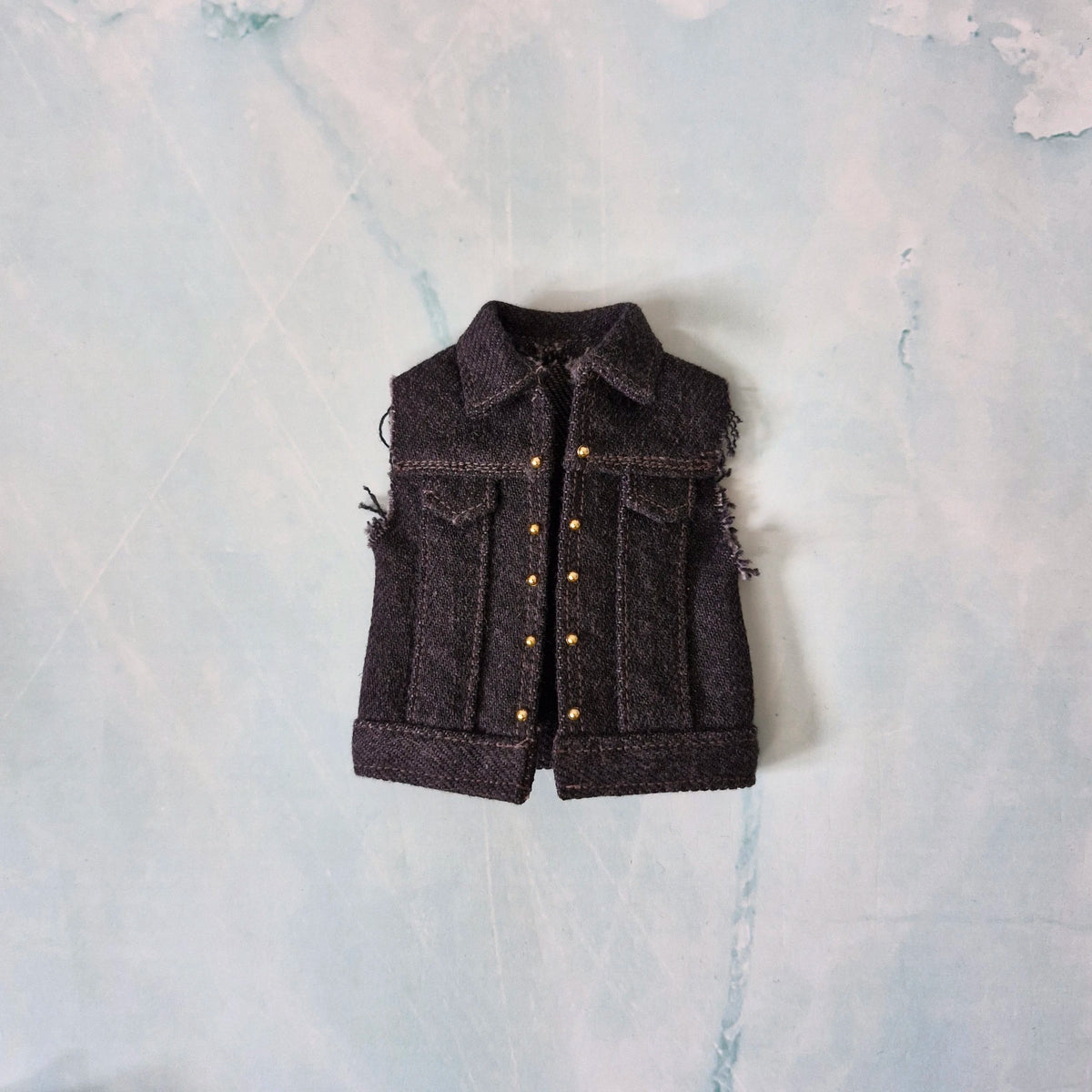 Flat lay of a custom handmade dark wash denim vest with gold stud details for Ken dolls.