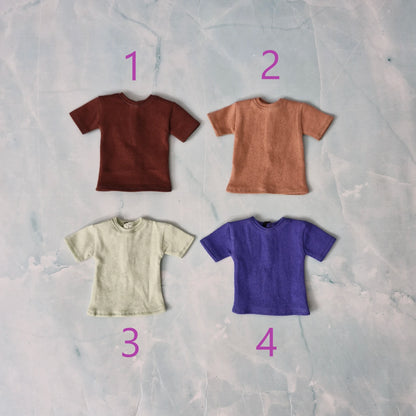 A flat lay image displaying multiple folded Ken doll t-shirts in blue, grey, and cream colors