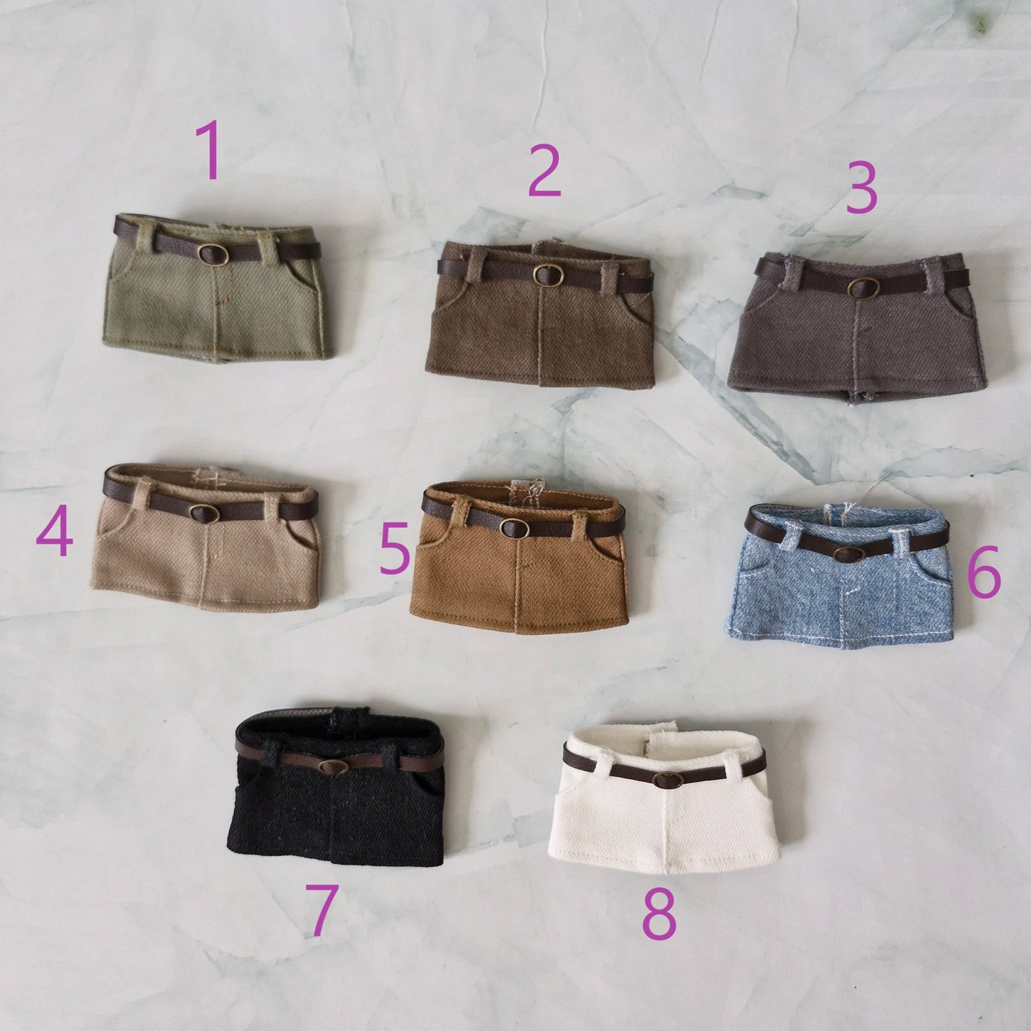 Eight miniature skirt accessories arranged on a light marble surface, numbered 1 through 8 in pink. The skirts come in olive green, brown, charcoal gray, beige, tan, light blue, black, cream, and brown leather-look belt details.