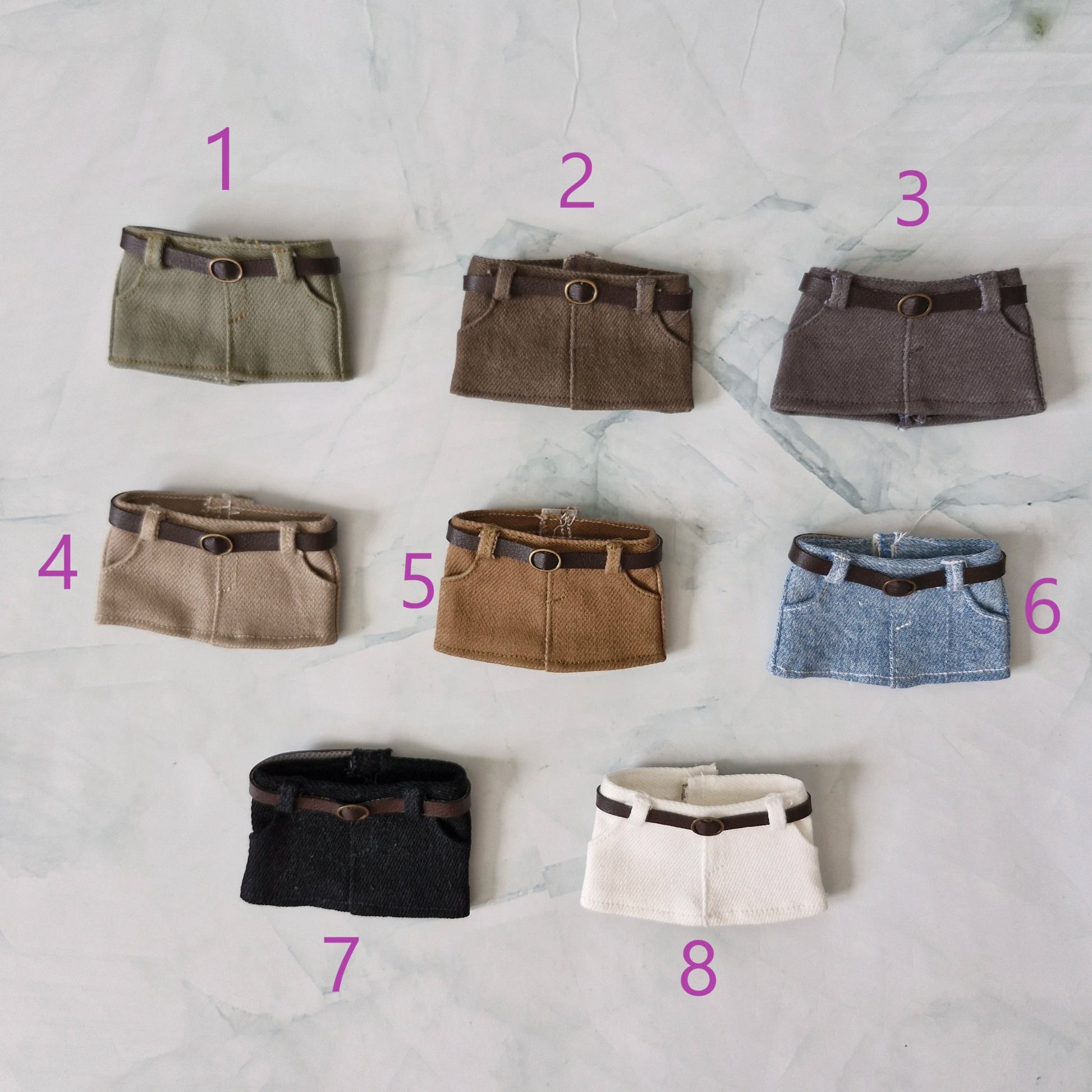 Eight miniature skirt accessories arranged on a light marble surface, numbered 1 through 8 in pink. The skirts come in olive green, brown, charcoal gray, beige, tan, light blue, black, cream, and brown leather-look belt details.