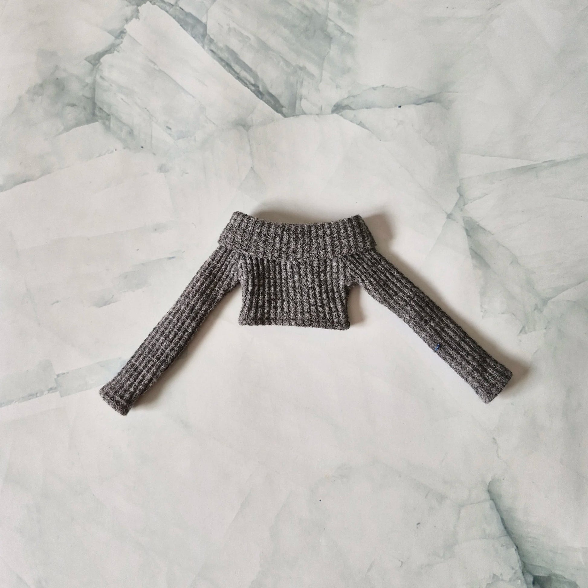 Flat lay of grey ribbed knit off-shoulder long-sleeve crop top sweater for fashion dolls on a marble background.

